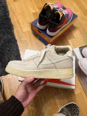 Nike Air Force 1 Stussy beige sneakers - Nike Air Force 1 Stussy sneakers i beige med low cut och platt sula. Ovandel i grovvävd canvas med broderad swoosh och Stussy-logga på tungan och hälen. Beige tonad sula och snörning framtill. Perfekt för dig som gillar clean och unik streetstil.