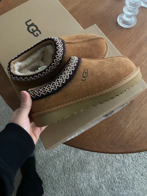 UGG tasman bruna mocka slip-ons med mönster - Säljer ett par bruna slip-on skor från UGG i mjuk mocka med fluffigt vitt foder. Skorna har en platt sula i beige och ett snyggt broderat mönster runt öppningen i svart och vitt. Perfekta för chill dagar hemma eller när du vill vara extra cozy.