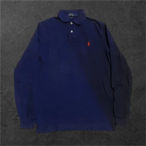 Marinblå långärmad pikétröja Polo Ralph Lauren - Snygg marinblå långärmad pikétröja från Polo Ralph Lauren med klassisk krage och två vita knappar. Tröjan har den ikoniska röda broderade loggan på bröstet och är tillverkad i mjuk bomull. Perfekt för dig som gillar stilrena och tidlösa plagg.