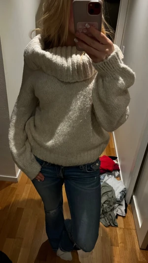 Beige offshoulder stickad tröja - Supermysig beige stickad tröja med bred ribbad krage som kan bäras offshoulder. Tröjan har långärmade, lite puffiga ärmar och en loose passform. Perfekt för att hålla sig varm och ändå se chill ut under hösten och vintern.