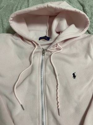 Ljusrosa hoodie från Polo Ralph Lauren - Snygg ljusrosa hoodie från Polo Ralph Lauren med dragkedja och huva. Tröjan har långa ärmar, snörning vid huvan och klassisk Polo-logga broderad på bröstet. Mjuk bomullskvalitet och praktiska fickor framtill. Perfekt för chill dagar.