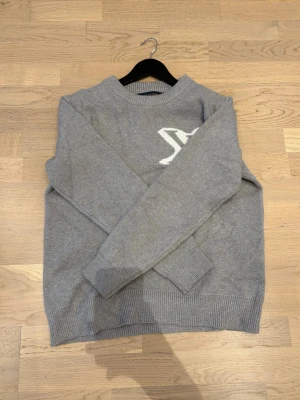 Grå stickad tröja Louis Vuitton - Snygg grå stickad tröja från Louis Vuitton med stort vitt LV-logo på bröstet. Tröjan har rund hals, långa ärmar och ribbade muddar vid ärmslut och nederkant. Perfekt för dig som gillar stilrena designer och exklusiva märken.