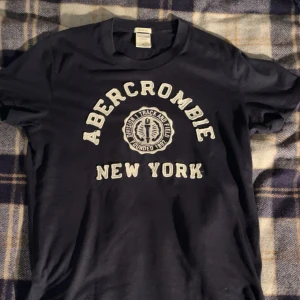 Blå Abercrombie t-shirt New York - Snygg blå t-shirt från Abercrombie & Fitch med vit broderad text 'Abercrombie New York' och loggan på bröstet. Perfekt för dig som gillar 2000: eran. 