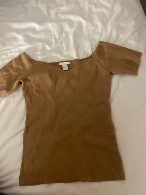 Beige offshoulder topp från H&M - Säljer en beige offshoulder topp från H&M i storlek S. Toppen har korta ärmar och en snygg v-ringad halsringning. Materialet är mjukt och stickat, vilket gör den både bekväm och stilren.