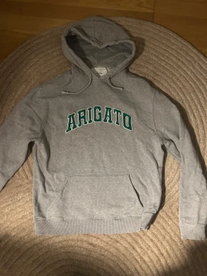 Grå hoodie Axel Arigato med grön text - Snygg grå hoodie från Axel Arigato i storlek M. Tröjan har en klassisk huva med snörning, magficka och grön text 'ARIGATO' på bröstet. 