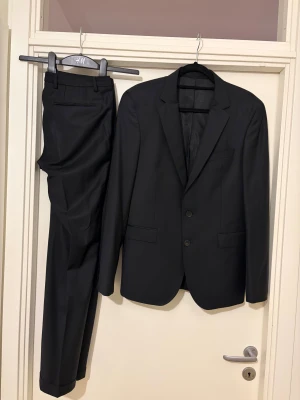 Svart kostym från Hugo Boss - Snygg svart kostym från Hugo Boss med klassisk kavaj och matchande kostymbyxor. Kavajen har två knappar, smala slag och innerfickor med stretchfoder. Stilren och tidlös design som ger en clean look. 