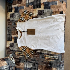 Vit Burberry t-shirt med rutiga ärmar - Vit t-shirt från Burberry med klassiska rutiga korta ärmar i beige, svart och rött. T-shirten har rund halsringning och är gjord i mjuk bomull. Perfekt för dig som vill ha en clean look med en touch av lyxig streetstyle.