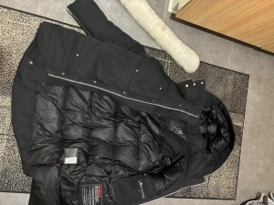 Svart dunjacka från Moose Knuckles - Säljer en svart lång dunjacka från Moose Knuckles med huva och snygga silverfärgade knappar. Jackan har ett quiltat innerfoder och den ikoniska loggan på bröstfickan. Perfekt för kalla dagar och har dragkedja samt tryckknappar framtill. Äkta med kvitto 