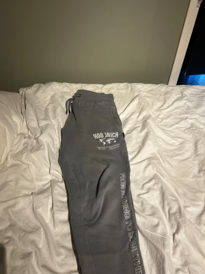 Grå Hoodrich mjukisbyxor med tryck - Snygga grå mjukisbyxor från Hoodrich med vit tryckt logga och världskarta på benet. Byxorna har snörning i midjan och texten 'FROM NOTHING TO SOMETHING' längs benet. Perfekt för chill eller streetwear. Materialet är mjukt och bekvämt bomull.