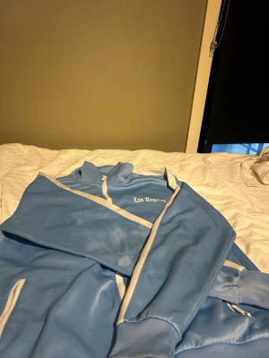 Blå vindjacka Black Squad S - Snygg ljusblå vindjacka från Black Squad med vita detaljer och dragkedja framtill. Jackan har ståkrage, vita ränder längs ärmarna och texten 'Los Angeles' tryckt på bröstet. Perfekt för dig som gillar sportig stil och streetwear. Med byxorna