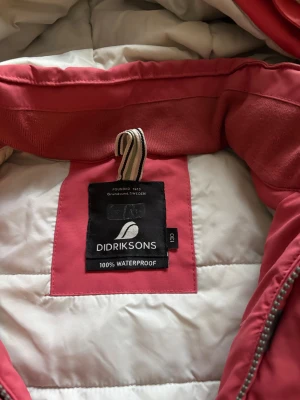 Rosa Didriksons vinterjacka med reflexpäls - Vinterjacka från Didriksons i en snygg ”rosa” färg med reflexdetaljer och fuskpäls och reflex på huvan. Jackan är 100% vattentät och har långa ärmar med kardborre vid mudden. Perfekt för kalla dagar och har praktiska fickor framtill. Dessvärre trasiga muddar som går att byta ut.