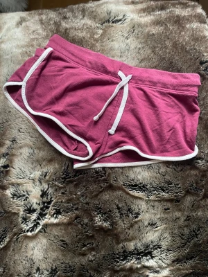 Röda sportiga shorts med vit kant - Säljer ett par snygga röda shorts med vit kant och snörning i midjan. De har en sportig look, rundade ben och en bakficka. Perfekta för chill dagar eller träning. Mjukt bomullsmaterial som känns skönt mot huden.