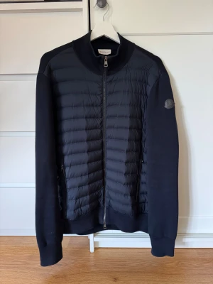 Svart quiltad pufferjacka Moncler L - Svart pufferjacka från Moncler i storlek L med quiltad front och släta ärmar. Jackan har två sidofickor med dragkedja, ribbad krage och Moncler-logga på vänster ärm. Materialmix med polyester och stickade detaljer. Snygg och stilren design med hel dragkedja framtill. Självklart äkta och jag har kvitto. Nypris 9900 kr, mitt pris 3800kr.