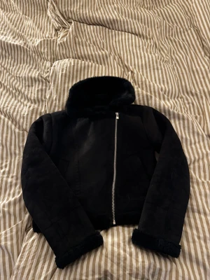 Svart pälsjacka -  svart pälsjacka med fluffig krage och muddar. Jackan har en snygg, rak passform och stängs med en silvrig dragkedja framtill. Utsidan är i mockaliknande material och insidan är fodrad med mjuk päls. Storlek L från Pull & Bear