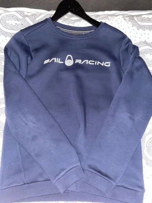 Marinblå Sail Racing sweatshirt - Snygg marinblå sweatshirt från Sail Racing med vit logotyp tryckt på bröstet och en mindre logga i nacken. Tröjan har rund hals, ribbade muddar och är långärmad. Perfekt för dig som gillar stilren design och sportiga märken.