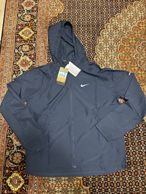 Marinblå vindjacka från Nike - Nike vindjacka i marinblått med huva och vit logga på bröstet. Jackan har dragkedja framtill, långa ärmar och diskreta fickor. Materialet är lätt och syntetiskt, perfekt för löpning eller andra sportaktiviteter. Snygg och sportig design med stilrena detaljer.