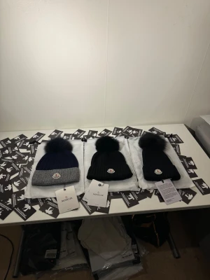 Moncler mössa med pälsboll - Snygg ribbstickad mössa från Moncler med stor fluffig pälsboll på toppen och klassisk Moncler-logga framtill. Finns i svart och mörkblå med grå kant. Perfekt för dig som vill ha en lyxig och trendig vinteraccessoar.