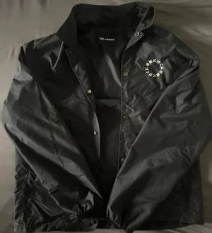 Axel Arigato - Limited Edition - Svart vindjacka från Axel Arigato i storlek M. Jackan har tryckknappar framtill och ett vitt tryck med japanska tecken på bröstet och på ryggen! Materialet är 55% polyamid och 45% polyester, vilket gör den lätt och smidig. Perfekt för dig som gillar minimalistisk streetstyle. Mycket bra skick behöver få sålt snabbt till studenten!