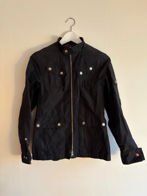 Svart vindjacka Peak Performance S - Svart vindjacka från Peak Performance med ståkrage, silverfärgade knappar och dragkedja framtill. Fyra fickor med lock och knappdetaljer, tillverkad i bomull och polyamid. Snygg och funktionell jacka med stilrena detaljer, perfekt för dig som gillar sportig look. Jackan är en äldre modell från Peak performance.