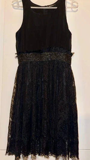 Svart långklänning med spets från Gina Tricot 36 - Elegant svart långklänning från Gina Tricot med ärmlös design, glittrigt midjeband och vacker spets i kjolen. Klänningen har rund halsringning och detaljerad spetskant nertill som ger en lyxig känsla. Perfekt för dig som vill sticka ut med stil.