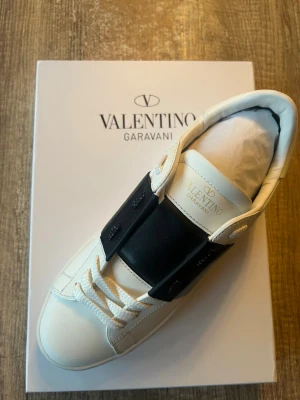 Valentino Garavani Open sneakers vit/svart - Äkta!!! Snygga Valentino Garavani Open sneakers i vitt skinn med bred svart läderdetalj över mitten. Skon har rund tå, platt sula med ikoniska nitar och vita snören. Märkeslogga på plösen och undersidan. Perfekt för dig som gillar stilrena designer med lyxig känsla. Utan box 3499