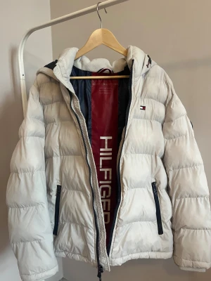 Vit pufferjacka från Tommy Hilfiger - Säljer en snygg vit pufferjacka från Tommy Hilfiger med luva och svarta dragkedjor. Jackan har broderad logga på bröstet och ett Hilfiger-märke på ärmen. Insidan är fodrad med röd och marinblå detalj. Perfekt för kalla dagar och riktigt stilren.