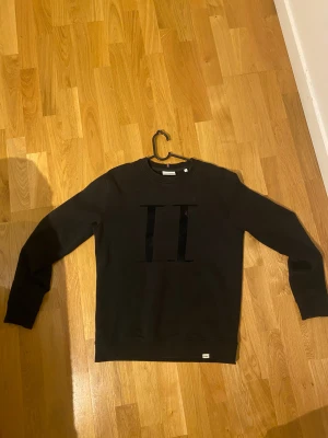 Svart sweatshirt Les Deux med tryck - Svart sweatshirt från Les Deux i storlek S. Tröjan har ett stort, svart sammetsliknande II-tryck på bröstet och ribbade muddar vid ärmslut och nederkant. Klassisk rund hals och långärmad modell, perfekt för en chill och stilren look.