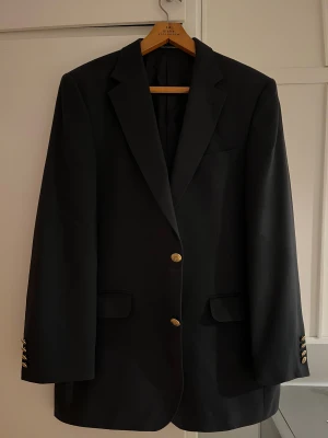 Mörkblå kavaj från Oscar Jacobson - Stilren mörkblå kavaj från Oscar Jacobson med snygga guldknappar framtill och på ärmarna. Klassisk passform med spetsiga slag och fickor med lock. Perfekt för dig som vill ha en tidlös och elegant look.