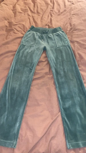 Gröna velourbyxor Juicy Couture XS - Snygga gröna mjukisbyxor från Juicy Couture i velour med snörning i midjan och raka ben. Byxorna har stora fickor fram och bak med knappdetaljer. Perfekta för en chill och trendig look.