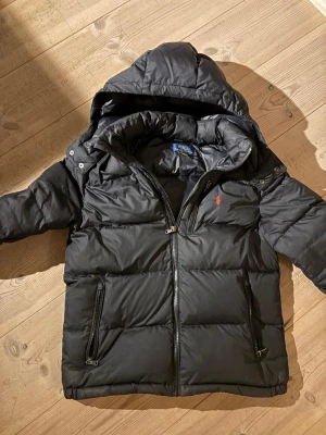 Svart pufferjacka från Polo Ralph Lauren - Svart pufferjacka från Polo Ralph Lauren med huva och dragkedja framtill. Jackan har två fickor med dragkedja och klassisk röd logga på bröstet. Perfekt för kalla dagar och har en stilren, sportig look.