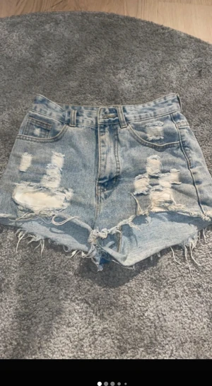 Ljusa denimshorts med slitningar - Säljer ett par ljusblå denimshorts med råa, fransiga kanter och stora slitningar framtill. Klassisk femficksmodell med knapp och dragkedja. Perfekta för dig som gillar en edgy och avslappnad stil.
