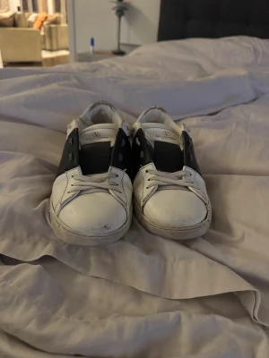 Valentino Garavani vita sneakers 40 - Säljer ett par snygga Valentino Garavani sneakers i vitt med bred svart läderdetalj över sidan. Klassisk rund tå, platt sula och vita snören. Märkeslogga på plösen och insidan. Perfekt för dig som gillar stilrena designer och exklusiva märken.