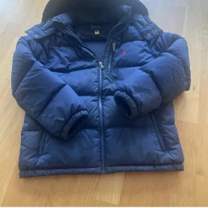 Mörkblå pufferjacka från Polo Ralph Lauren - Diskuterar gärna pris                                                                                Säljer en mörkblå pufferjacka från Polo Ralph Lauren med huva och broderad röd logga på bröstet. Jackan har två sidofickor med dragkedja och RL-detaljer på zipparna. Perfekt för kalla dagar 