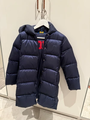 Mörkblå dunjacka Polo Ralph Lauren - Säljer en lång, mörkblå dunjacka från Polo Ralph Lauren med huva och stora, quiltade sektioner. Jackan har dragkedja och tryckknappar framtill samt röd POLO-logga på insidan. Perfekt för kalla dagar och riktigt snygg streetstil.