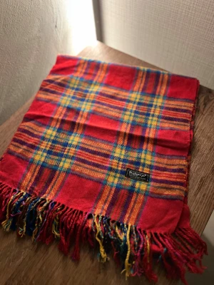 Rutig röd halsduk från Burberrys - Klassisk rutig halsduk från Burberrys i 100% ull. Halsduken har fransar i ändarna och den ikoniska Burberrys-etiketten. Perfekt accessoar för att lyfta din outfit. Anledningen att de stör ”burberrys” är för att de är en äldre modell.