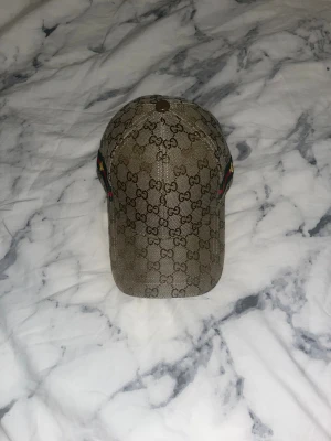 Gucci keps beige med brodyr - Snygg beige keps från Gucci med klassiskt GG-monogram över hela ytan. På sidan finns en broderad gul bi på en grön och röd rand. Insidan är svart med Gucci-logga på banden. Justerbar rem baktill. Materialet är canvas och bomull.