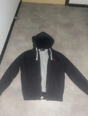 Mörkgrå hoodie Polo Ralph Lauren - Snygg mörkgrå hoodie från Polo Ralph Lauren med röd broderad logga på bröstet. Tröjan har huva med vita snören, två fickor fram och dragkedja. Mjuk insida i fleece och långärmad passform som är normal. Perfekt för chill dagar och streetwear.