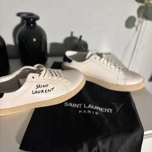 Saint Laurent vita sneakers i skinn - Snygga vita sneakers från Saint Laurent med handskriven logga på sidan. Skorna har rund tå, platt beige sula och svarta detaljer vid hälen. Tillverkade i slätt skinn med klassisk snörning och Saint Laurent Paris tryckt på insidan och undersidan av sulan.