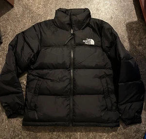 Svart pufferjacka från The North Face - Säljer en svart pufferjacka från The North Face med hög krage och tydlig logga på bröstet och ryggen. Jackan har dragkedja framtill, två sidofickor med dragkedja och elastiska muddar vid ärmslut. Perfekt för kalla dagar och riktigt snygg streetstil.