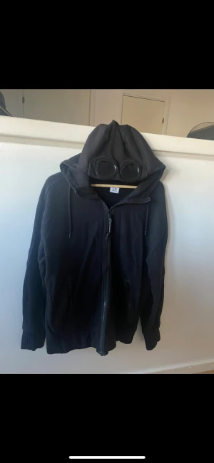 C.P. Company Zip Hoodie - Säljer en riktigt snygg svart C.P. Company hoodie med goggles-detalj på huvan. Storlek M och 100% äkta. Den är använd men i bra skick och har alltid förvarats noggrant. Först till kvarn är det som gäller! Skicka dm vid frågor eller fler bilder😊