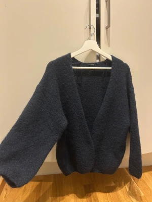 Mörkblå stickad kofta från Lindex - Mysig och oversized mörkblå stickad kofta från Lindex med djup v-ringning och långa ärmar. Perfekt för att slänga över en t-shirt eller linne när det är lite kyligt. Koftan har en fluffig struktur och känns riktigt skön mot huden.