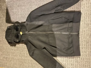 Svart hoodiejacka från C.P. Company - Svart hoodiejacka från C.P. Company med ikoniska goggles i huvan och broderad logga på bröstet. Jackan har två stora fickor framtill och hel dragkedja. Materialet är mjukt och känns som bomull, perfekt för en chill streetwear-look.