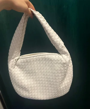 Vit axelväska i flätad skinn från NA-KD - Snygg vit axelväska från NA-KD med flätad design och mjukt syntetskinn. Väskan har en bred axelrem och stängs med dragkedja. Perfekt storlek för det viktigaste❤️‍🔥❤️‍🔥