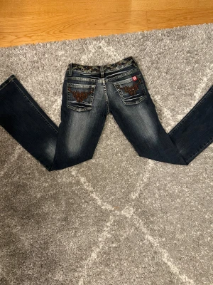 M.K Jeans blå bootcut med broderi - Snygga mörkblå bootcut jeans från M.K Jeans med slitningar framtill och detaljerad broderad design på bakfickorna. Midjan har ett mönstrat band och passformen är normal. Jeansen är tillverkade i klassiskt denim och har en cool, ungdomlig vibe.