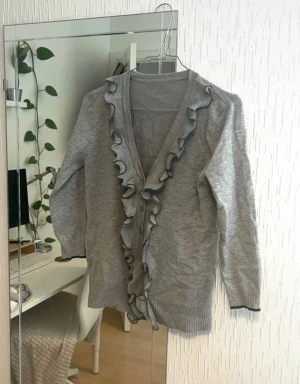 Grå kofta med volangdetaljer - Säljer en grå kofta med snygga volangdetaljer längs v-ringningen och knappar framtill. Koftan har lång ärm och ribbade muddar vid ärmslut och nederkant. Materialet är mjuk bomull och polyester, perfekt för lager-på-lager under kyliga dagar. Som ny!