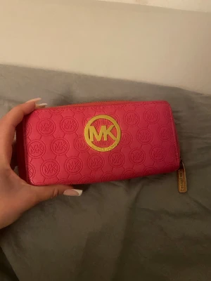 Rosa MK plånbok med gulddetaljer - Säljer en snygg rosa plånbok från Michael Kors med guldfärgat MK-emblem och dragkedja. Plånboken har präglat MK-mönster över hela ytan och flera fack för kort och mynt. Perfekt storlek för att ha med sig överallt.