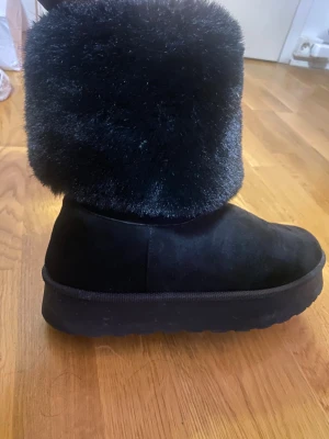 Svarta fluffiga boots med platåsula - Säljer ett par svarta boots med hög fluffig päls upptill och platåsula. Skorna har rund tå och är tillverkade i mocka och syntetpäls, perfekta för kalla dagar när du vill hålla stilen. Riktigt mysiga och trendiga för vintern.