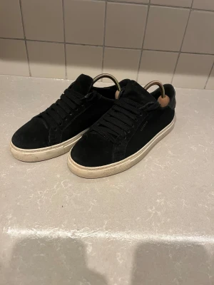 Svarta sneakers från Axel Arigato - Snygga svarta sneakers från Axel Arigato i mocka med klassisk vit sula. Skorna har rund tå, platt sula och svart snörning. Perfekta för dig som gillar stilrena och minimalistiska skor med premiumkänsla.