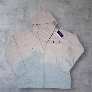 Grå hoodie från Polo Ralph Lauren - Snygg ljusgrå hoodie från Polo Ralph Lauren med dragkedja och huva. Klassisk Polo-logga broderad i svart på bröstet. Mjuk bomullsblandning, ribbade muddar och fickor framtill. Perfekt för chill dagar och streetwear-stil.