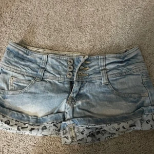 Ljusblåa jeansshorts från RSK - Säljer ett par ljusblåa denimshorts från RSK med låg midja och snygga detaljer. Shortsen har tre knappar framtill, uppvikta benslut med svart tryck och klassiska fickor både fram och bak. Perfekta för sommaren och riktigt trendiga.